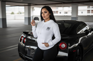 Woman wearing white M Line hoodie holding car-shaped air freshener in front of black Nissan GTR — Silhouette Collection by M Line Automotive.